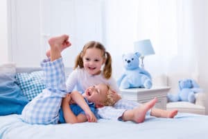 Happy kids playing in white bedroom. Little boy and girl, brother and sister play on the bed wearing pajamas. Nursery interior for children. Nightwear and bedding for baby and toddler. Family at home.